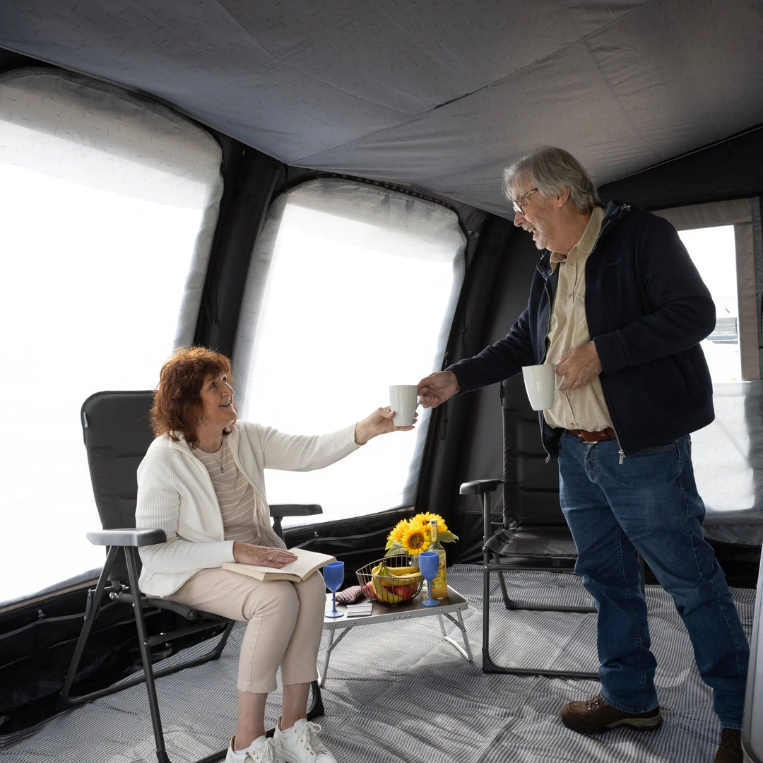 A woman sits in a black camping chair inside a Vango Tuscany Pro Air 400 Caravan Awning, reaching to share a white mug with an older man standing beside her. A small folding table holds a flower arrangement and blue glasses.