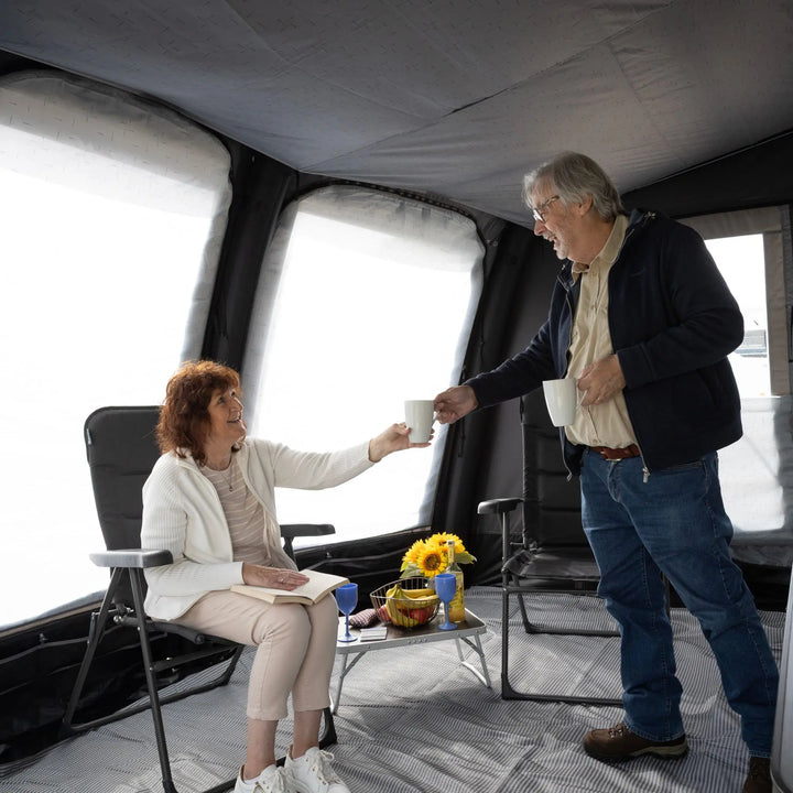 A woman sits in a black camping chair inside a Vango Tuscany Pro Air 400 Caravan Awning, reaching to share a white mug with an older man standing beside her. A small folding table holds a flower arrangement and blue glasses.