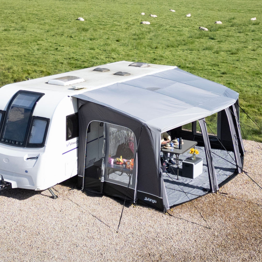 A Vango Tuscany Pro Air 500 Caravan Awning set up on a gravel campsite, with a green grassy field and sheep in the background. A person is visible sitting inside the awning area near a small table.