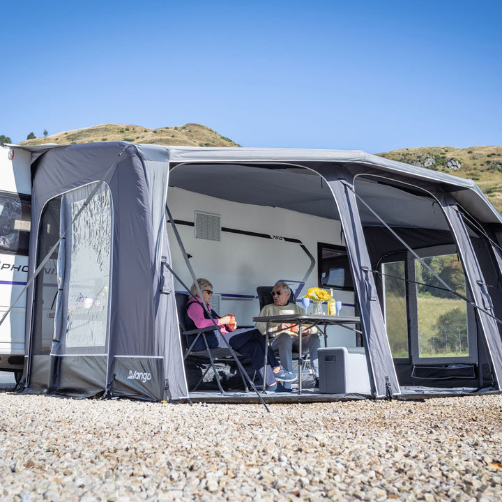 A Vango Tuscany Pro Air 500 Caravan Awning is pitched over a white caravan on a gravel campsite with two people sitting inside under a blue sky and hills in the background.