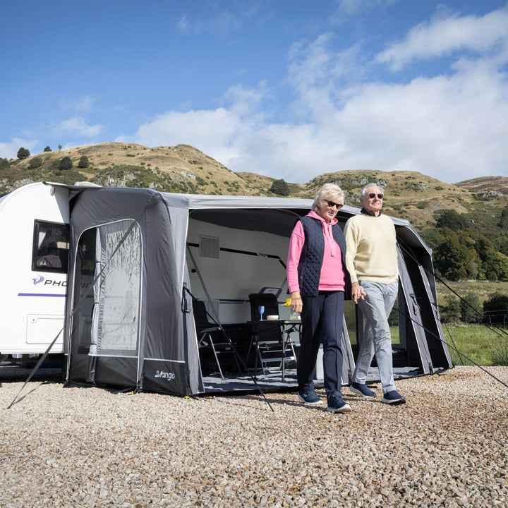A Vango Tuscany Pro Air 500 Caravan Awning pitched beside a white caravan on a gravel surface, with two people standing in front. Rolling hills and blue sky are visible in the background.