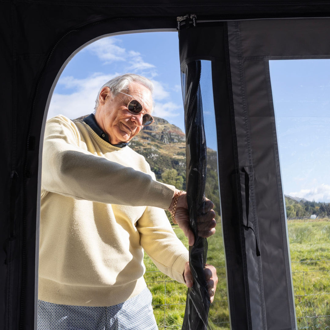 An older man wearing sunglasses and a cream sweater stands at the open doorway of a Vango Tuscany Pro Air 500 Caravan Awning, holding the black zipper panel to close it. A grassy field and a mountain are visible in the background under a blue sky.