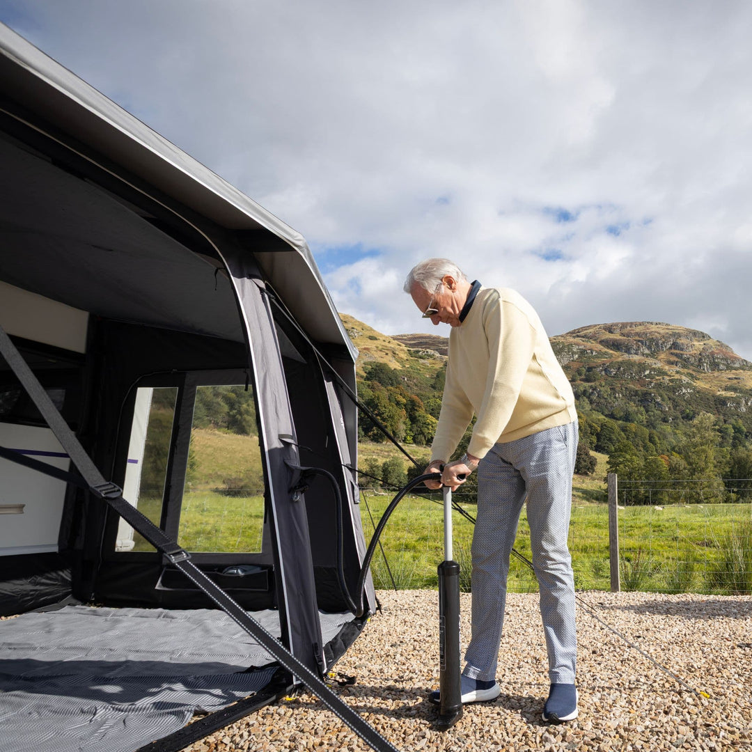 An older man pumps air into a Vango Tuscany Pro Air 500 Caravan Awning extension outdoors on a gravel surface, with a grassy field and hills under a cloudy sky in the background.