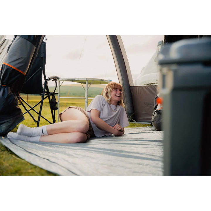 Woman relaxing inside the Vango Versos Air Mid drive-away awning, lying on the sewn-in groundsheet with a scenic outdoor view.