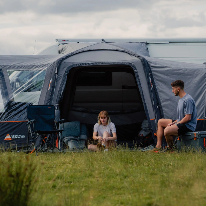 A woman sitting on the grass and a man on a storage box, unwinding in front of the Vango Versos Air Mid inflatable campervan awning. Highlighting its easy access fold flat door.