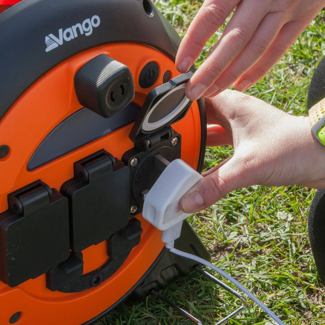 Person connecting a mains plug to the Vango Voltaic Roll Away Mains Unit on a grassy surface.