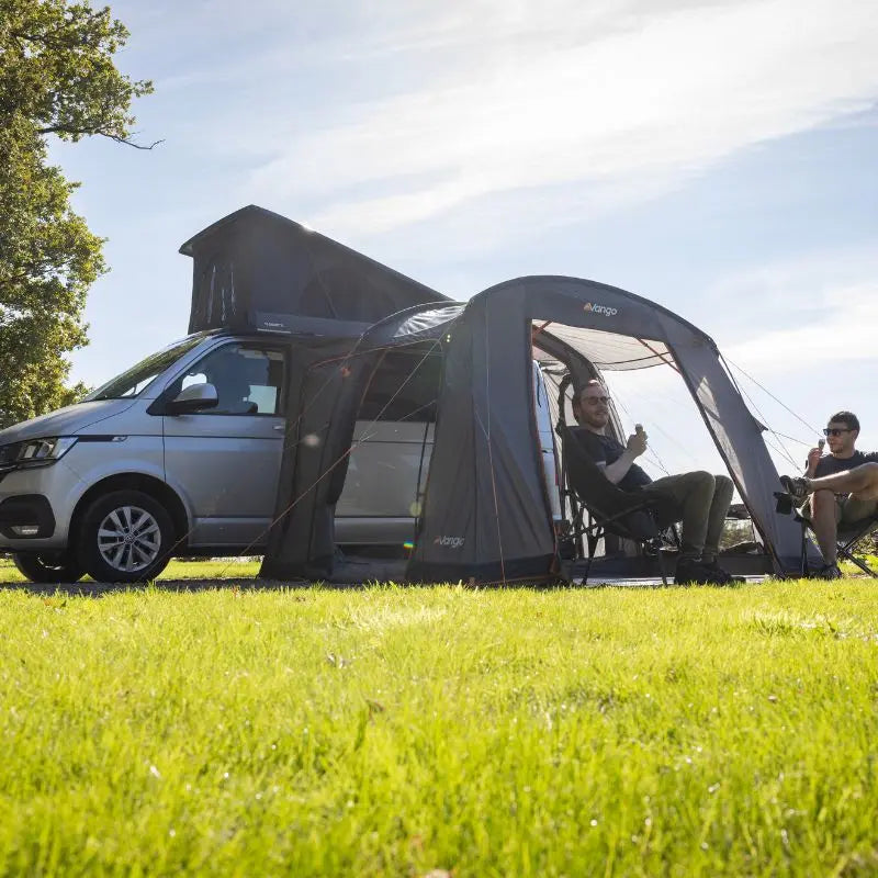 Two people sitting outside a VW Camper with an awning attached on a grassy area.