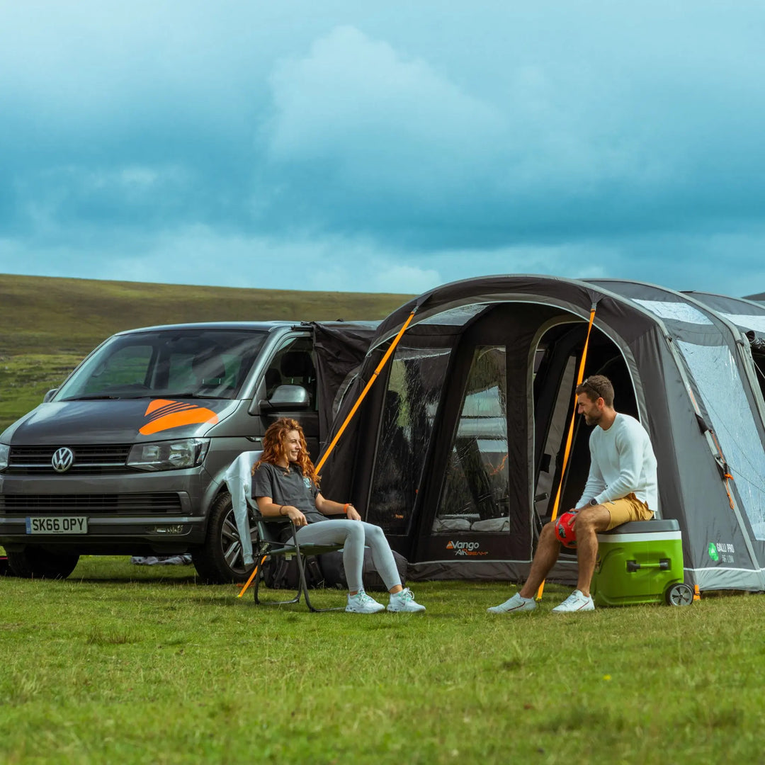 A grey Volkswagen campervan with an attached Vango drive-away awning set up on a grassy field, with a man and woman sitting outside enjoying the outdoors.